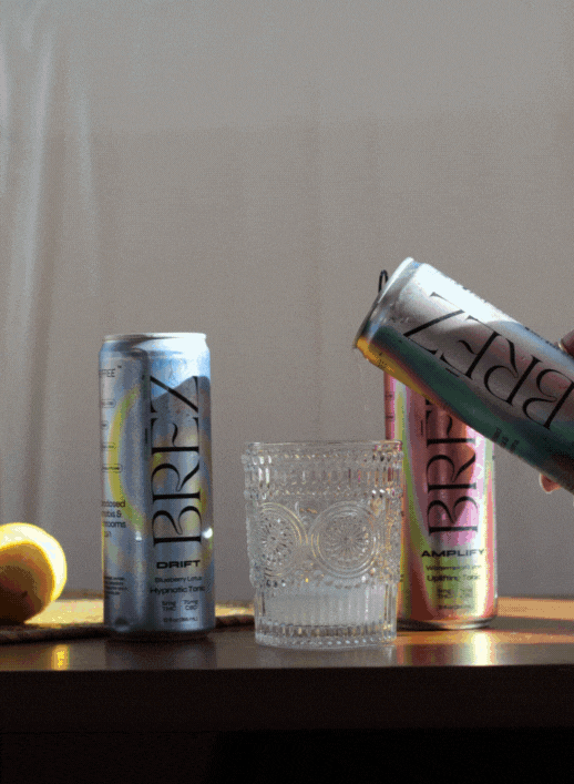 A hand pours a drink from a BRĒZ can into a patterned glass, with two other BRĒZ cans and a sliced lemon on a wooden surface in soft lighting.