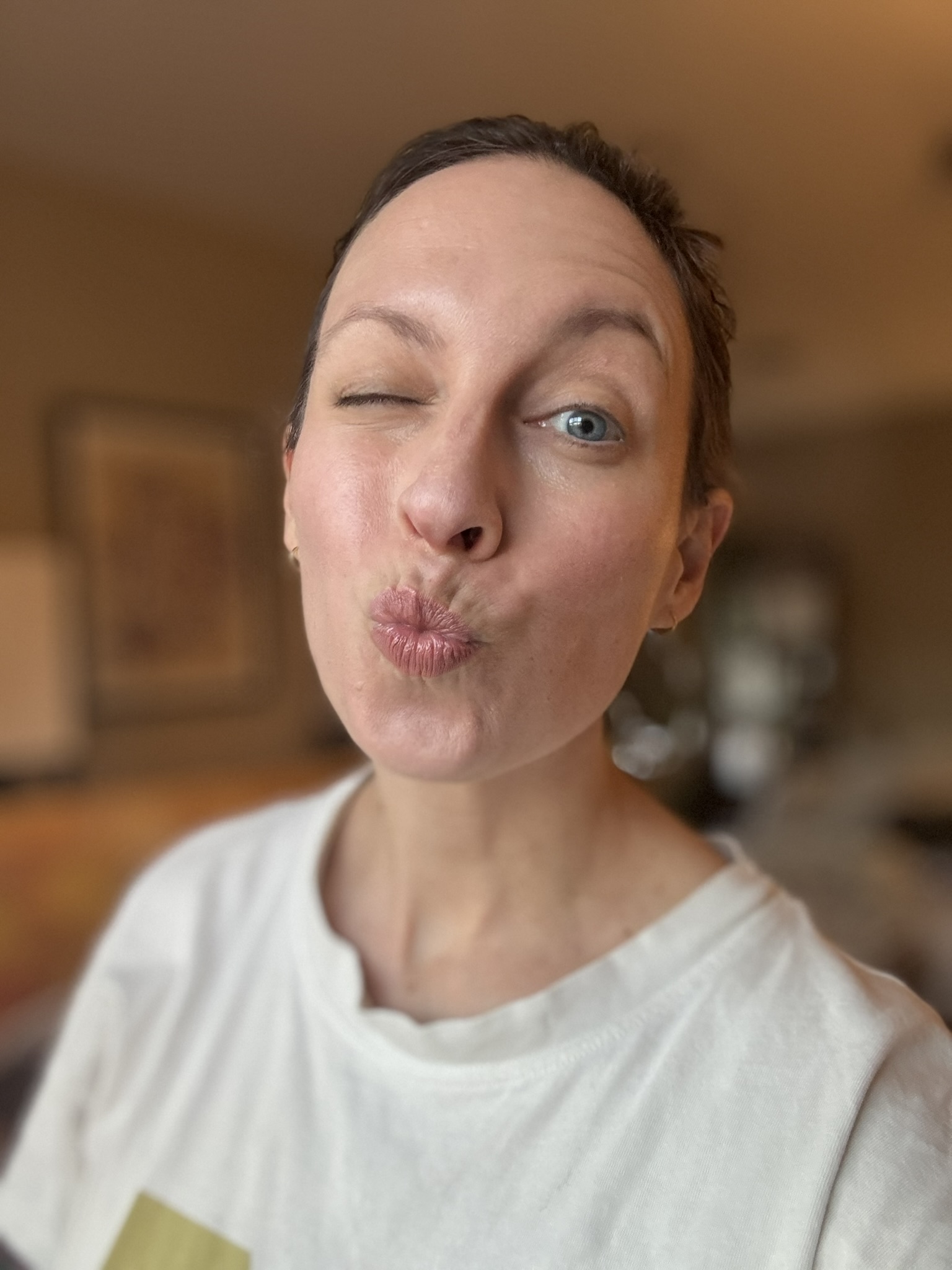 A person wearing a white shirt is winking with one eye and making a kissy face, standing indoors with a blurred background.
