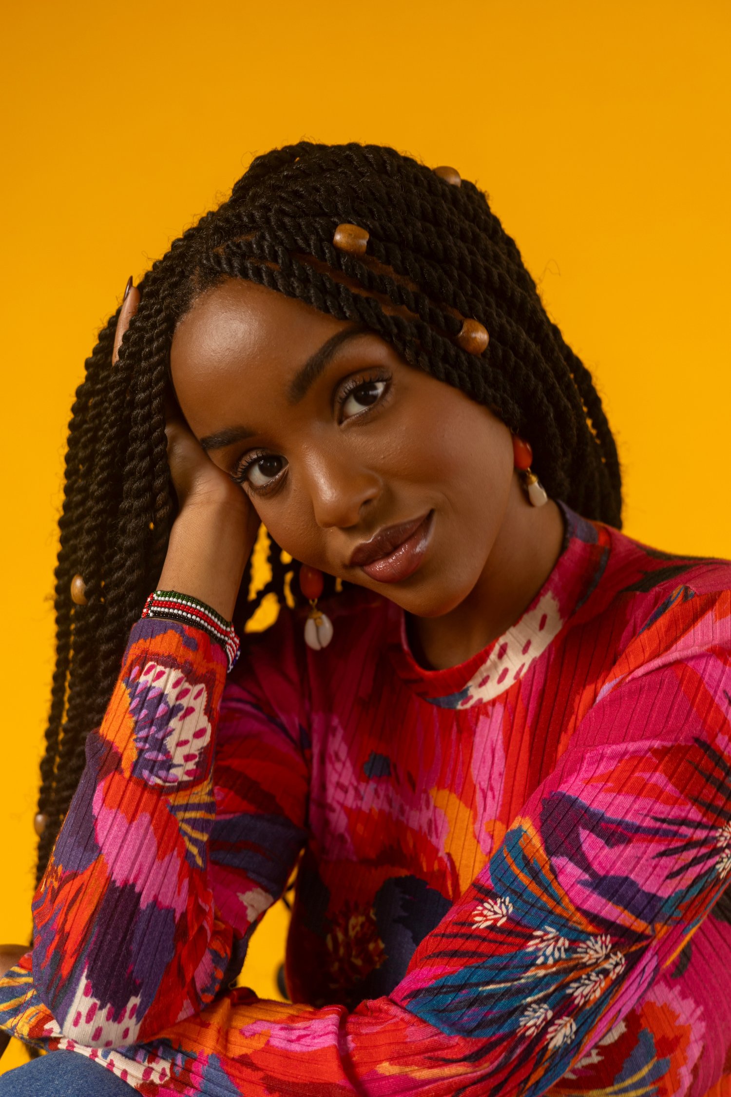 A woman with long, twisted braids adorned with beads rests her head on her hand, wearing a colorful floral top, against a solid yellow background.