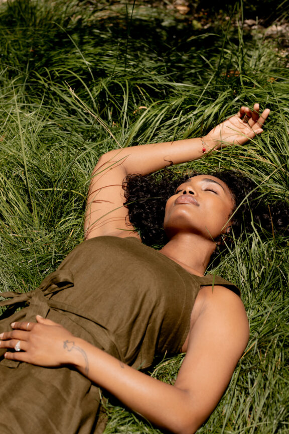 A woman in an olive green dress lies on her back in tall grass with her eyes closed, one hand resting on her stomach and the other extended above her head.