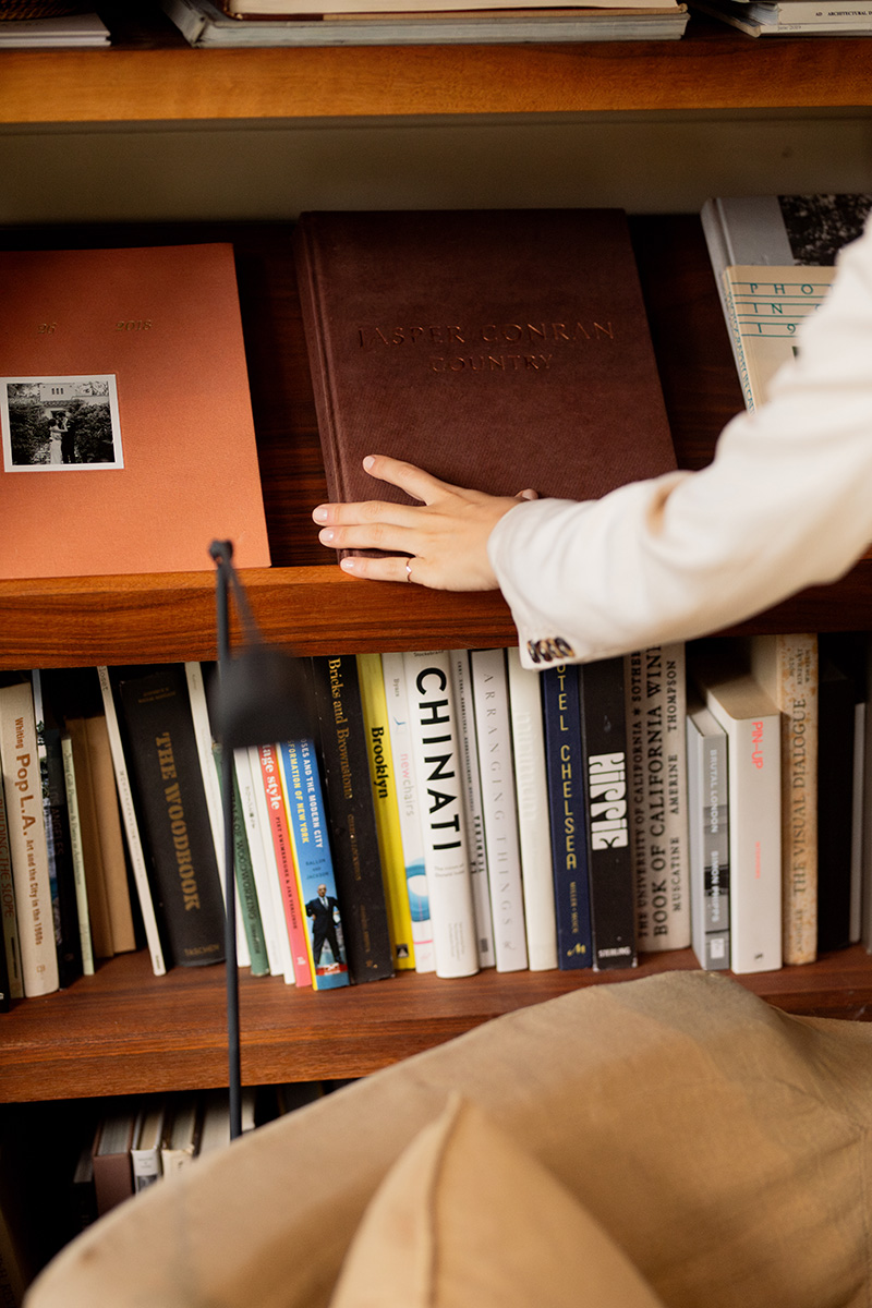 A person’s hand reaches for a large brown book on a wooden bookshelf filled with various books and a reading lamp in the foreground.