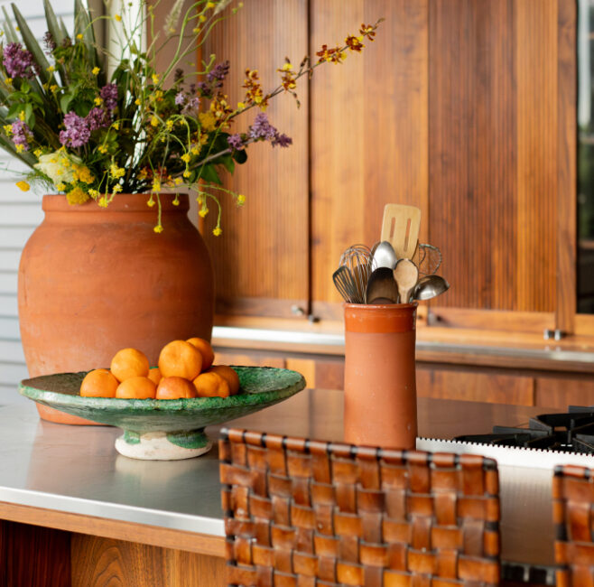 A kitchen counter with a woven chair, a large vase of flowers, a bowl of peaches, and a ceramic holder with cooking utensils.