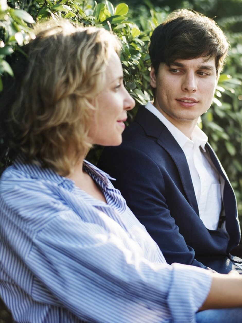 Two people sitting on a bench outdoors, engaged in conversation. The person on the left holds a take-away coffee cup. Green foliage is visible in the background.