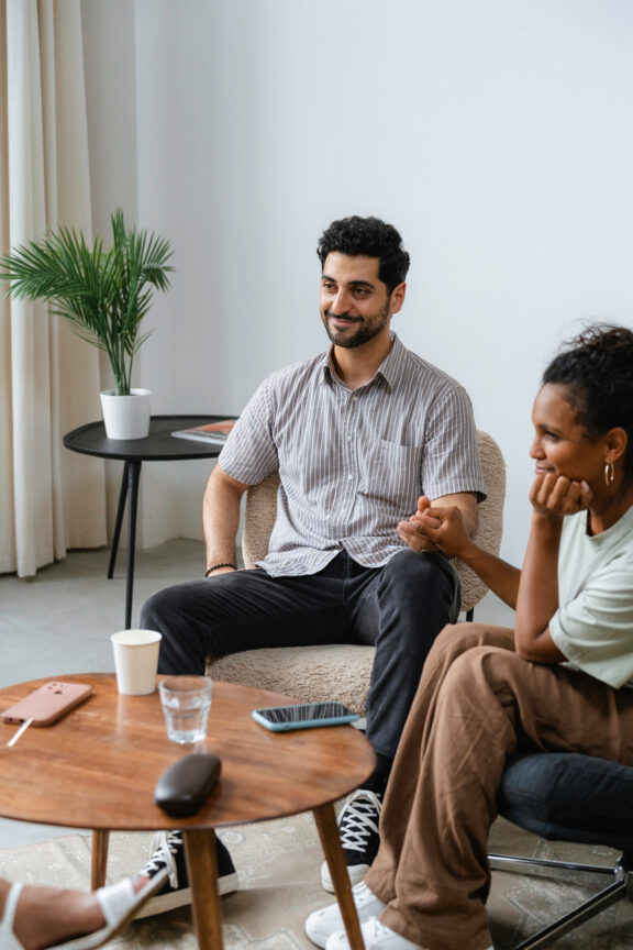 A man and a woman sitting in a living room, engaged in conversation. A small coffee table with a smartphone, glass, and cup is in front of them, and a potted plant is in the background.