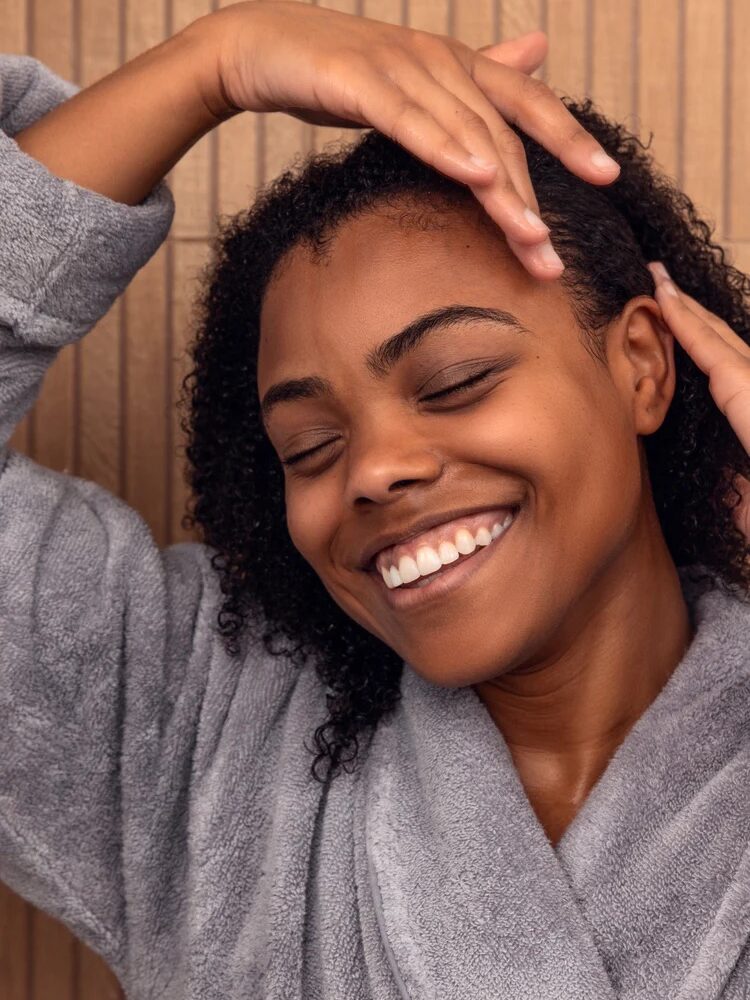 A joyful woman smiling with her eyes closed, hands on her head, wearing a gray robe.