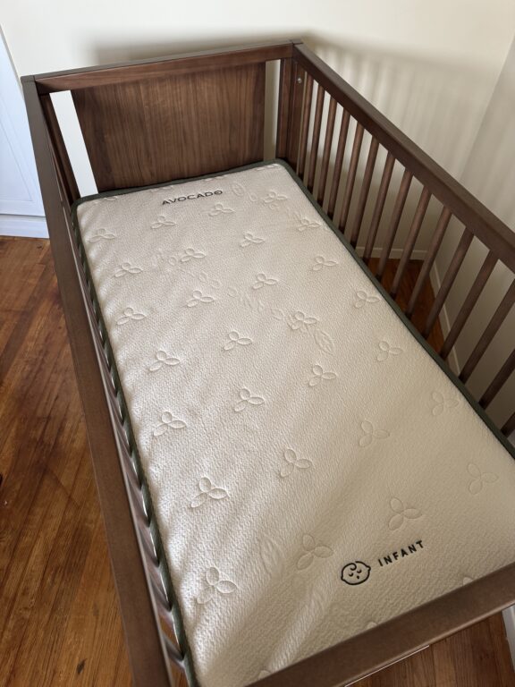 A wooden baby crib with a white Avocado brand infant mattress inside, placed on a hardwood floor.