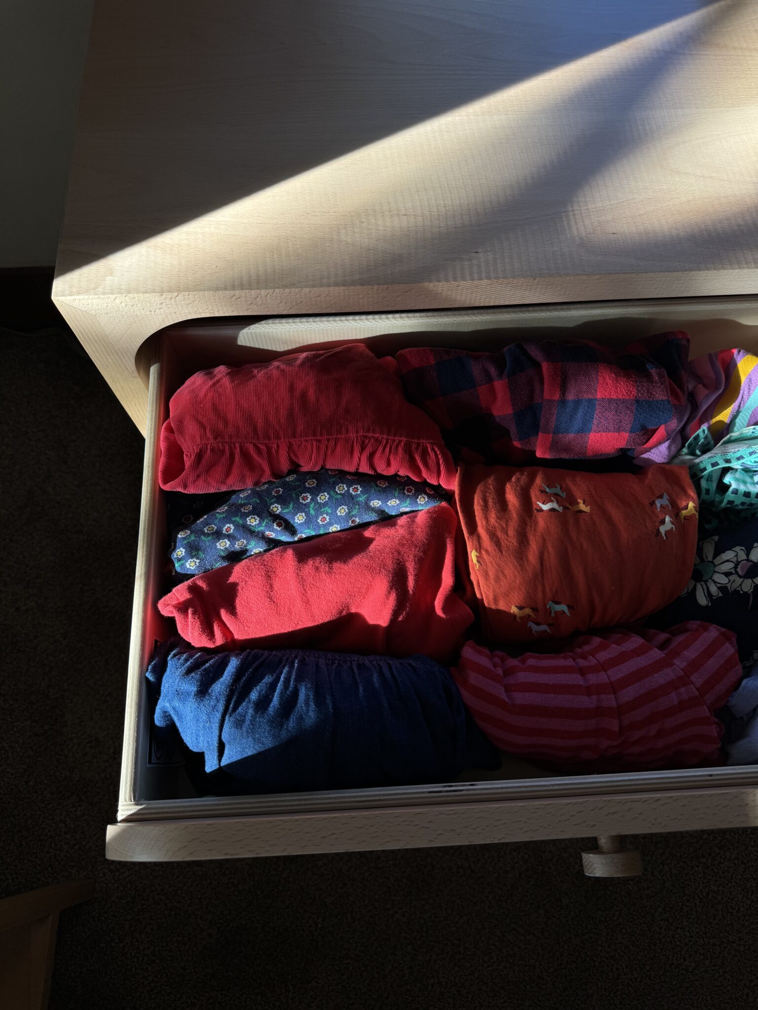 An open wooden drawer holds neatly folded clothes in various patterns and colors, including red, blue, and plaid, lit by sunlight from the side.