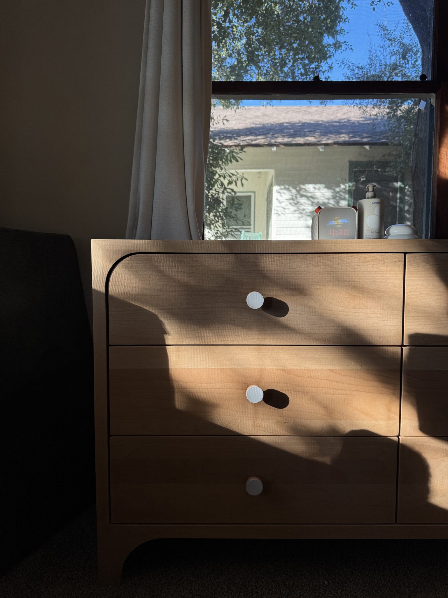 A wooden dresser with three drawers is partially in shadow, positioned in front of a window showing a sunlit house and trees outside. Several small items sit on top of the dresser.