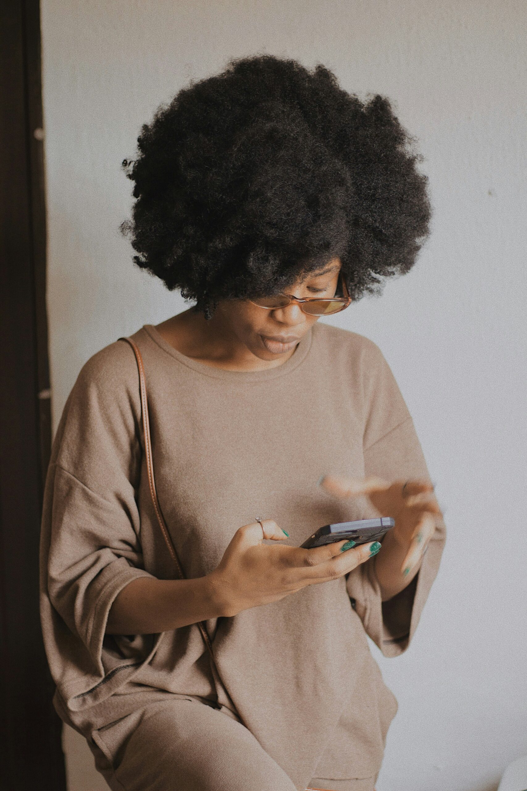 A person with curly hair and glasses is wearing a loose, beige outfit and using a smartphone while standing indoors against a plain, light-colored wall.