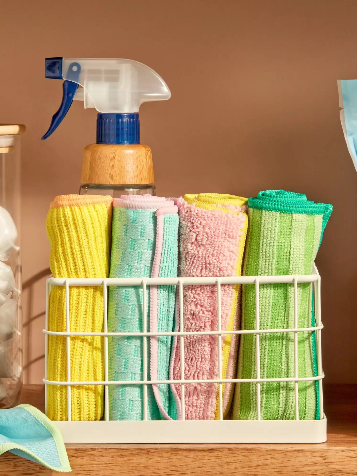 A white wire basket holds neatly folded microfiber cloths in pastel colors beside a spray bottle on a wooden surface.