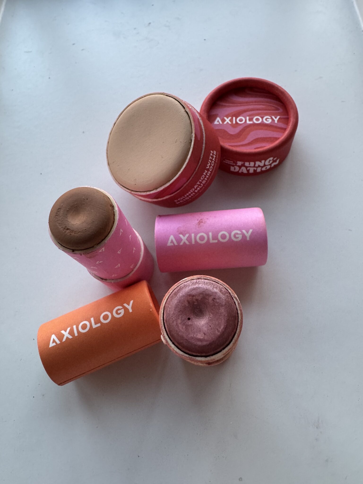 Three Axiology makeup sticks in cardboard tubes, each opened to show different product shades in beige, brown, and mauve, placed on a white surface.