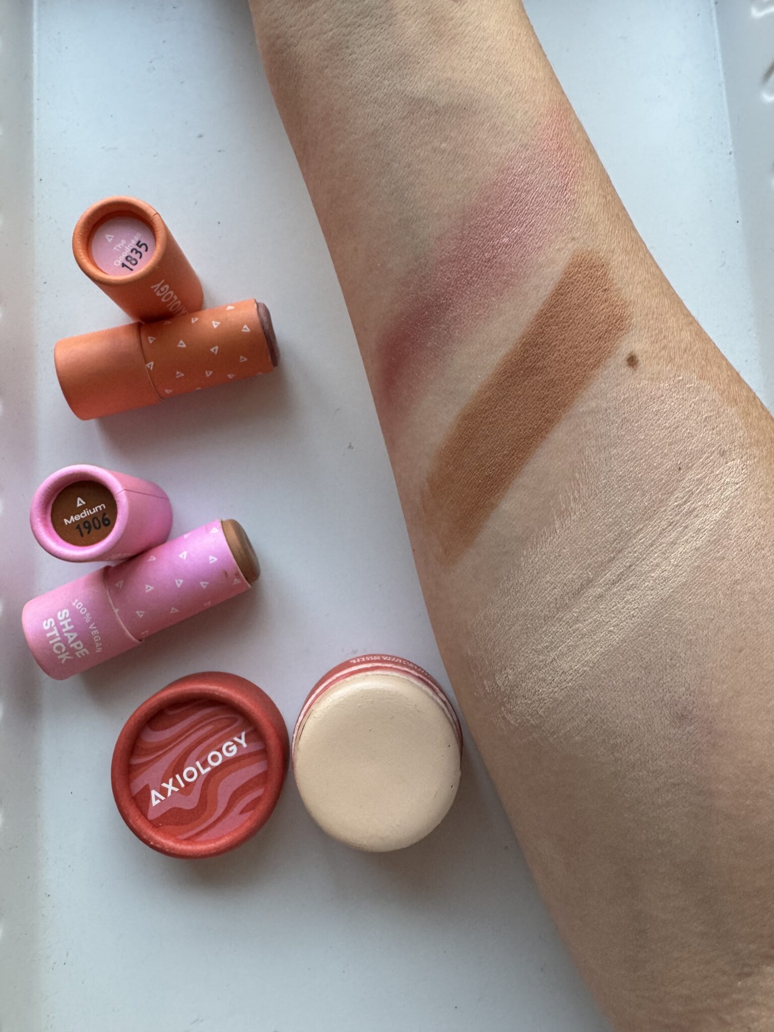An arm with swatches of makeup in pink, brown, and shimmery shades is shown next to four tubes of lipstick and an open blush compact labeled "Axiology" on a white surface.
