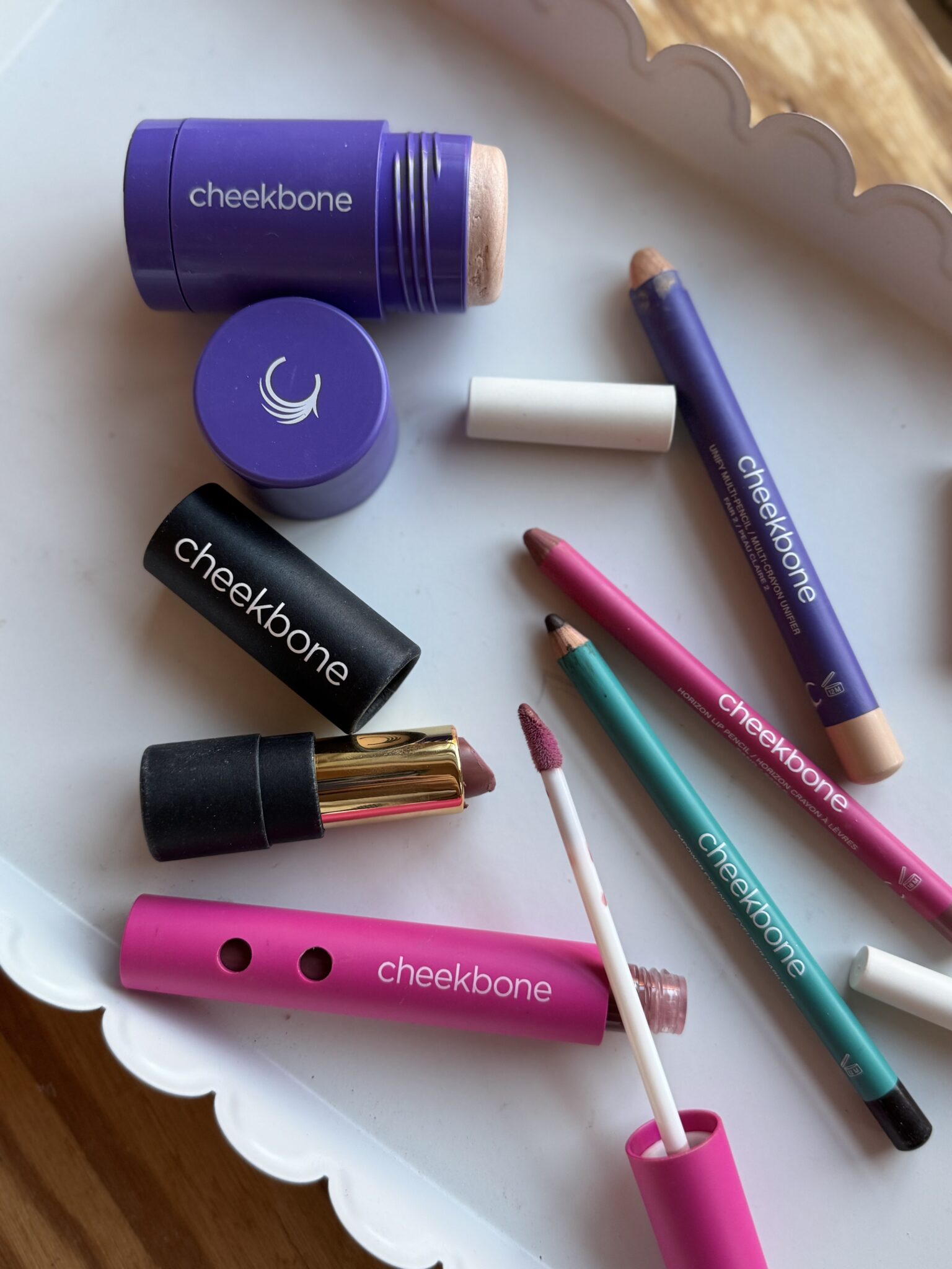 Various Cheekbone brand makeup products, including lipstick, lip gloss, lip pencil, eyeshadow crayon, and highlighter, arranged on a white tray.