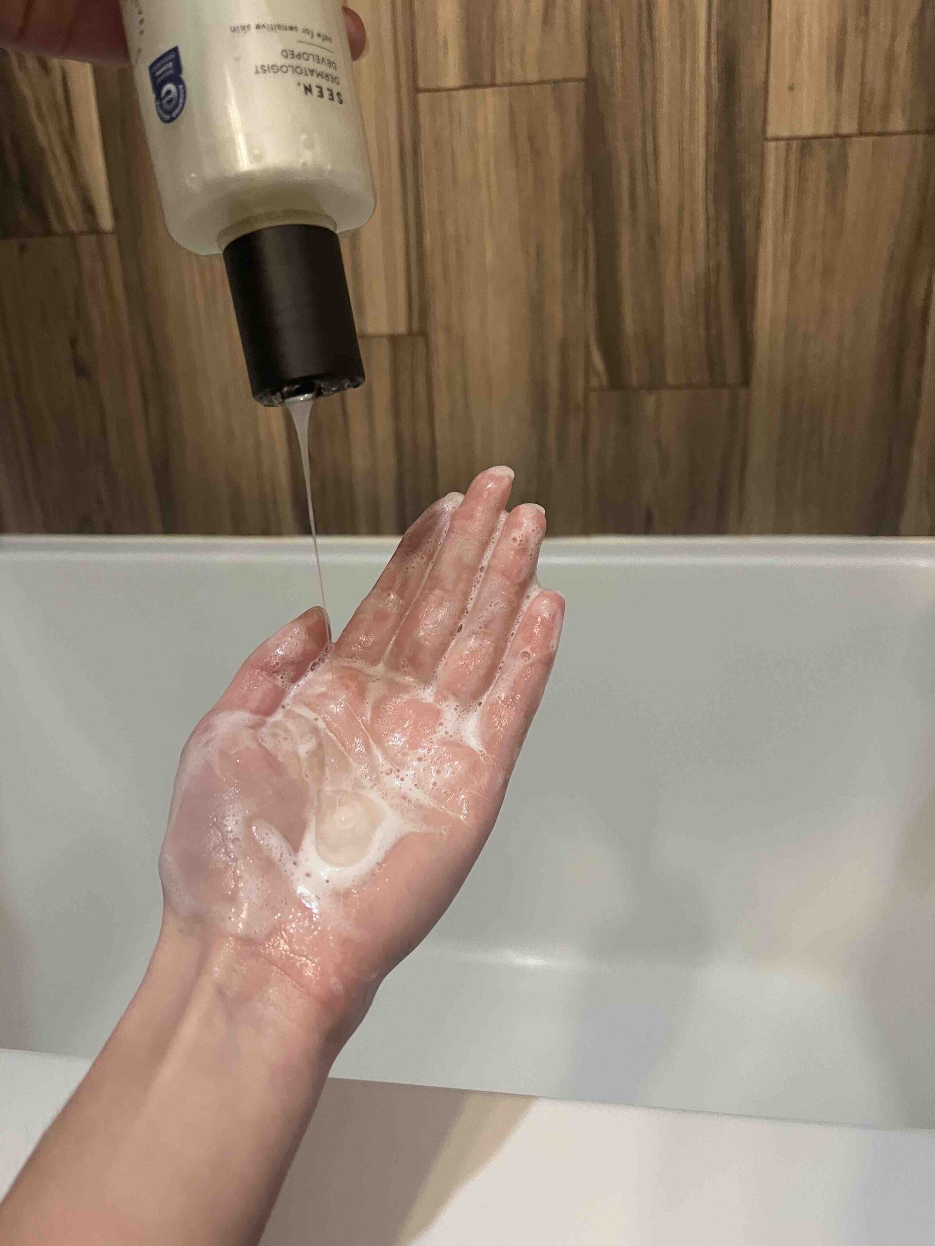 Person dispensing liquid soap onto a lathered hand in a bathroom with wood-paneled walls.