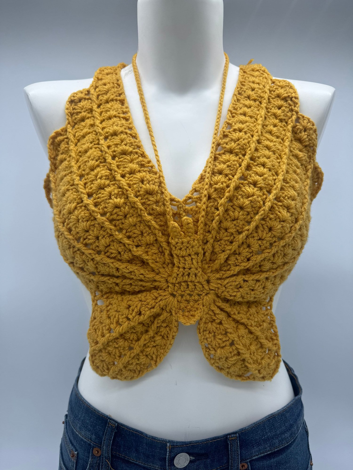 Crocheted mustard yellow halter top with a butterfly pattern on a mannequin, paired with blue jeans.