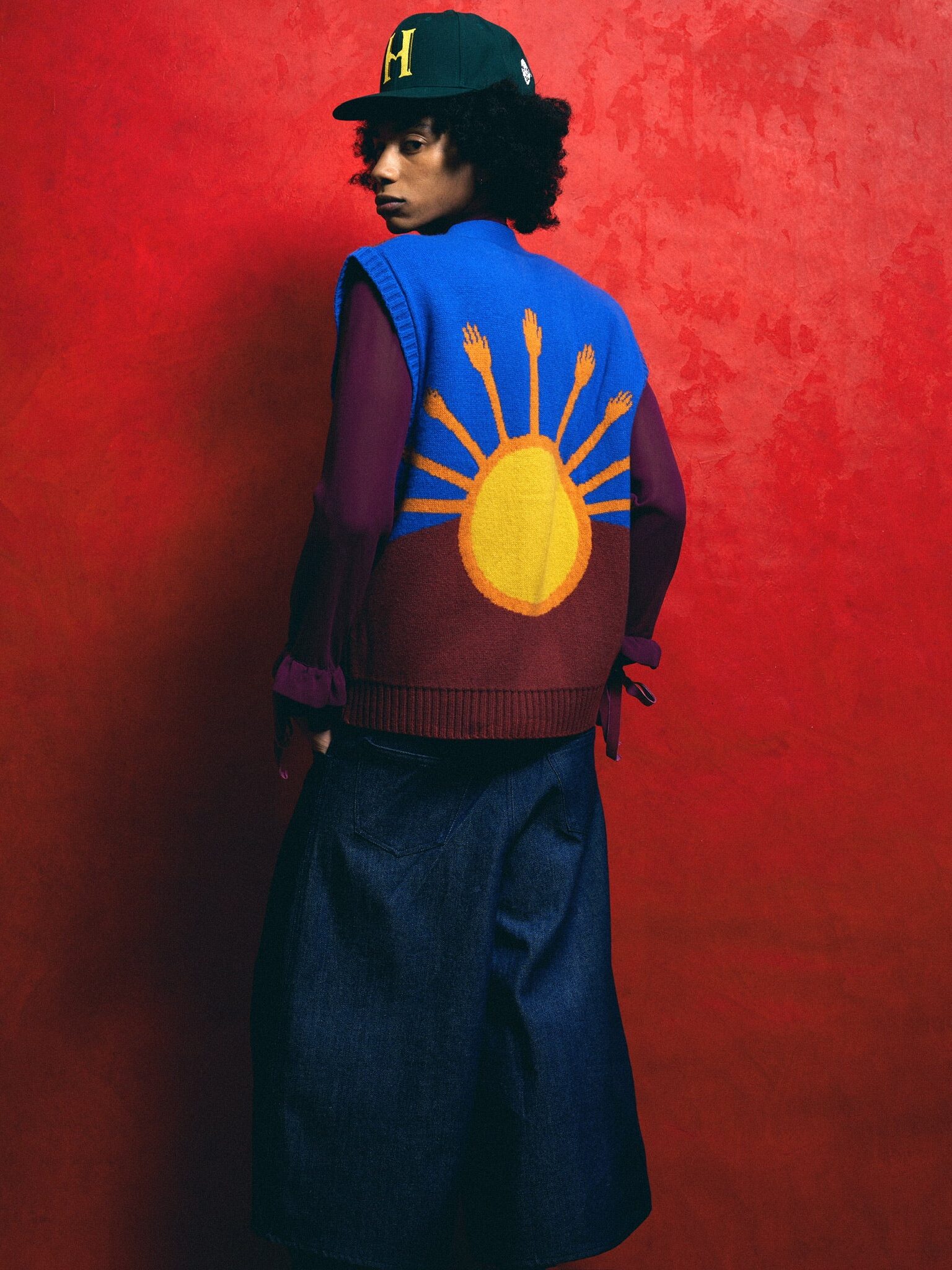 Person wearing a colorful vest with a sun design, wide denim pants, and a green cap, standing against a red textured background.