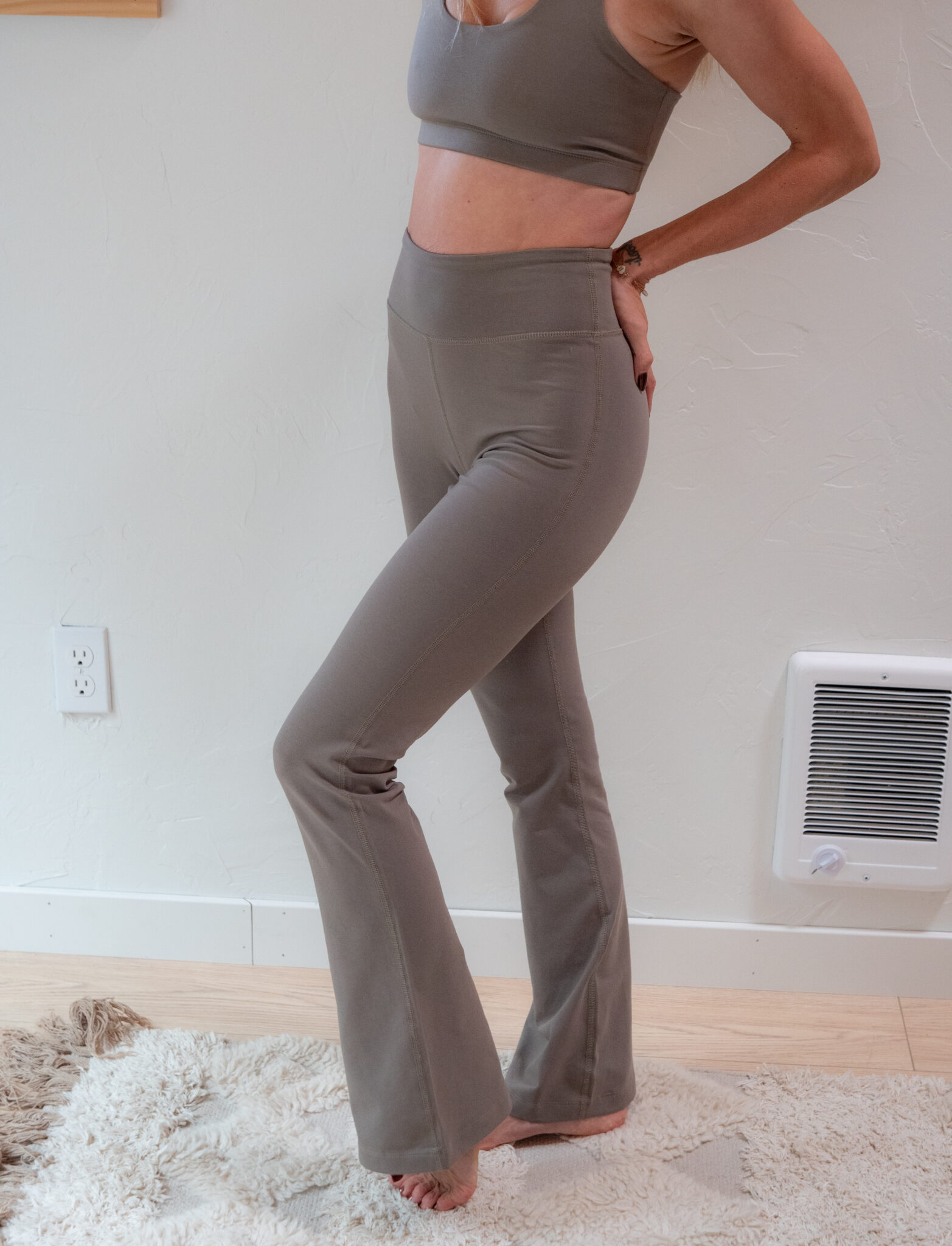 Person wearing a matching gray sports bra and high-waisted pants standing barefoot on a rug in a room with light-colored walls and a heater on the wall.