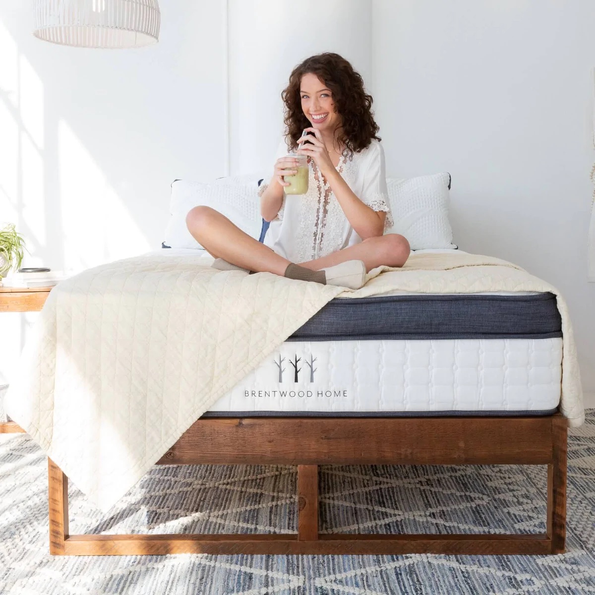 A person sits cross-legged on a bed with a quilt, savoring a drink, in a bright room. The bed frame reads "Brentwood Home," showcasing an organic mattress known for its nontoxic qualities.