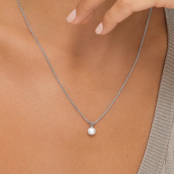 A close-up of a person wearing a silver chain necklace with a single round pearl pendant, shown against bare skin and part of a beige top.