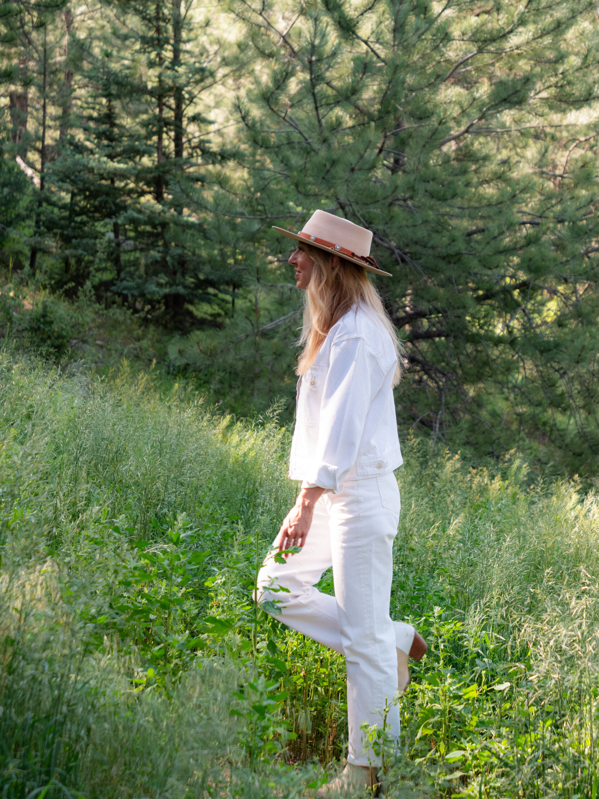 A woman dressed in white with a tan hat stands in a grassy field surrounded by trees on a sunny day.