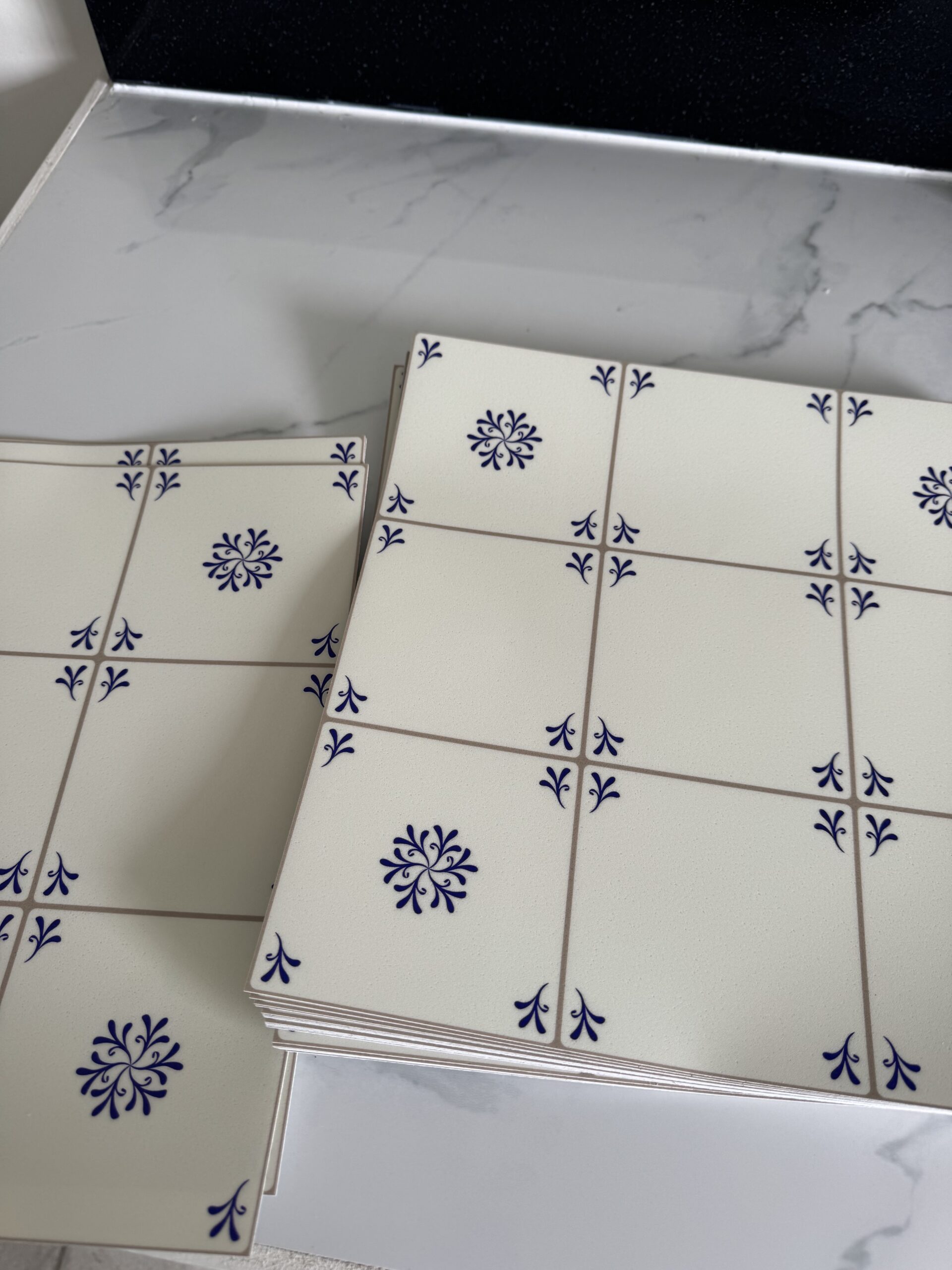 Two stacks of white ceramic tiles with blue floral and corner designs are placed on a white marble surface.
