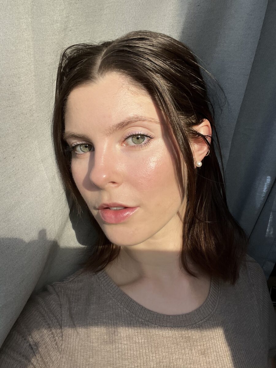 Person with dark hair, wearing a pearl earring, poses in front of a gray curtain with sunlight casting shadows across the face.