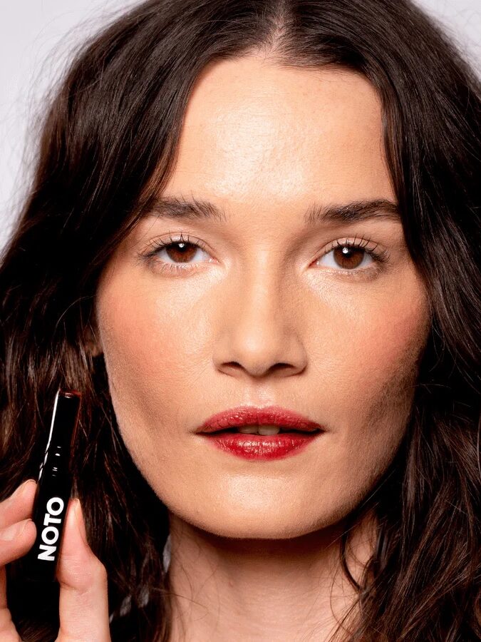 Person with long dark hair holding a lipstick tube labeled "NOTO" near their face, wearing red lipstick, against a plain background.