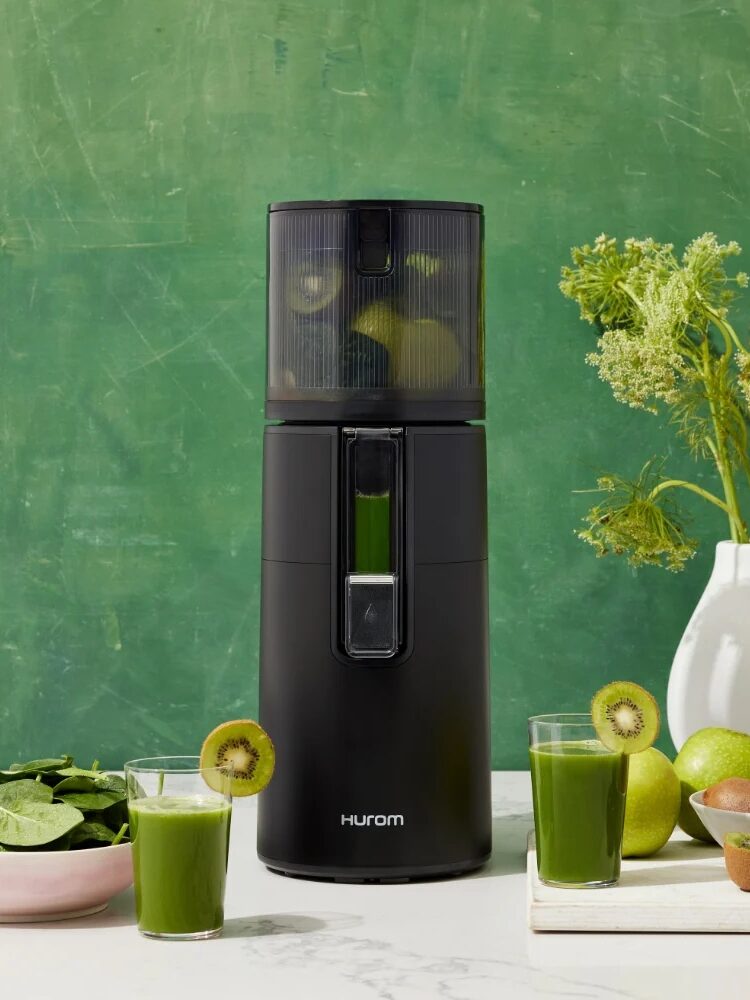A black Hurom juicer with green juice inside, surrounded by a glass of green juice, spinach, kiwis, and a plant, against a green textured background.