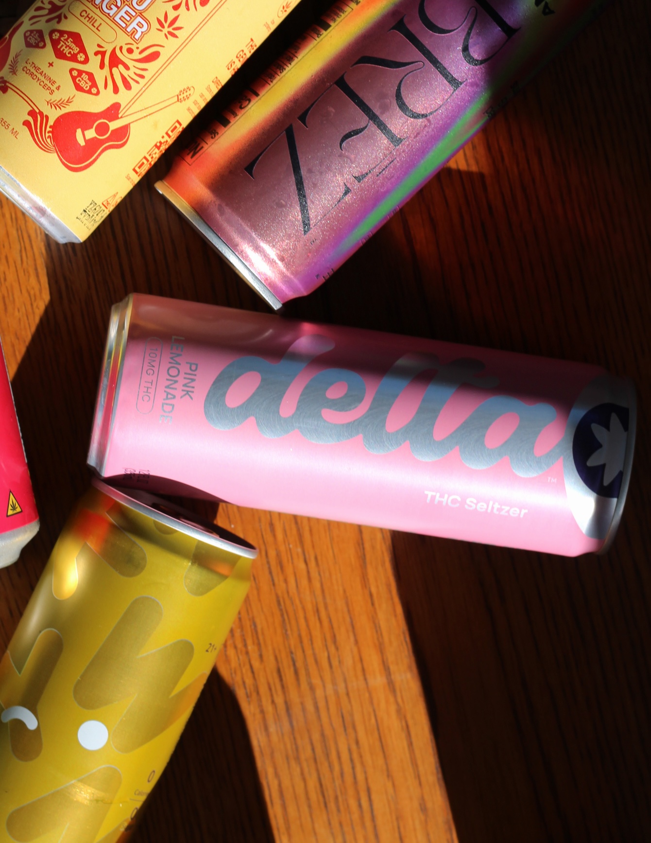 Several colorful beverage cans, including a pink "delta" THC seltzer can labeled "Pink Lemonade," are scattered on a wooden surface in natural light.