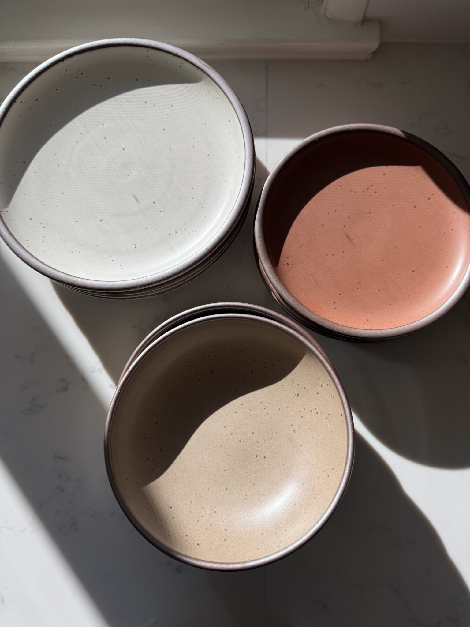 Three ceramic bowls in white, terracotta, and beige are arranged on a sunlit white surface, with shadows partially covering them.