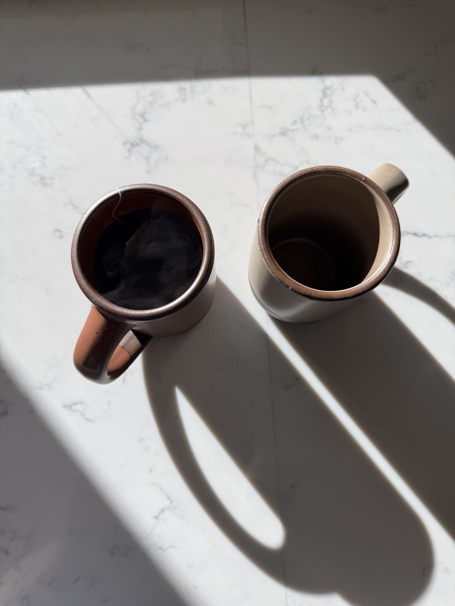 Two ceramic mugs on a white marble surface in sunlight, one filled with a dark liquid and the other empty, casting long shadows.