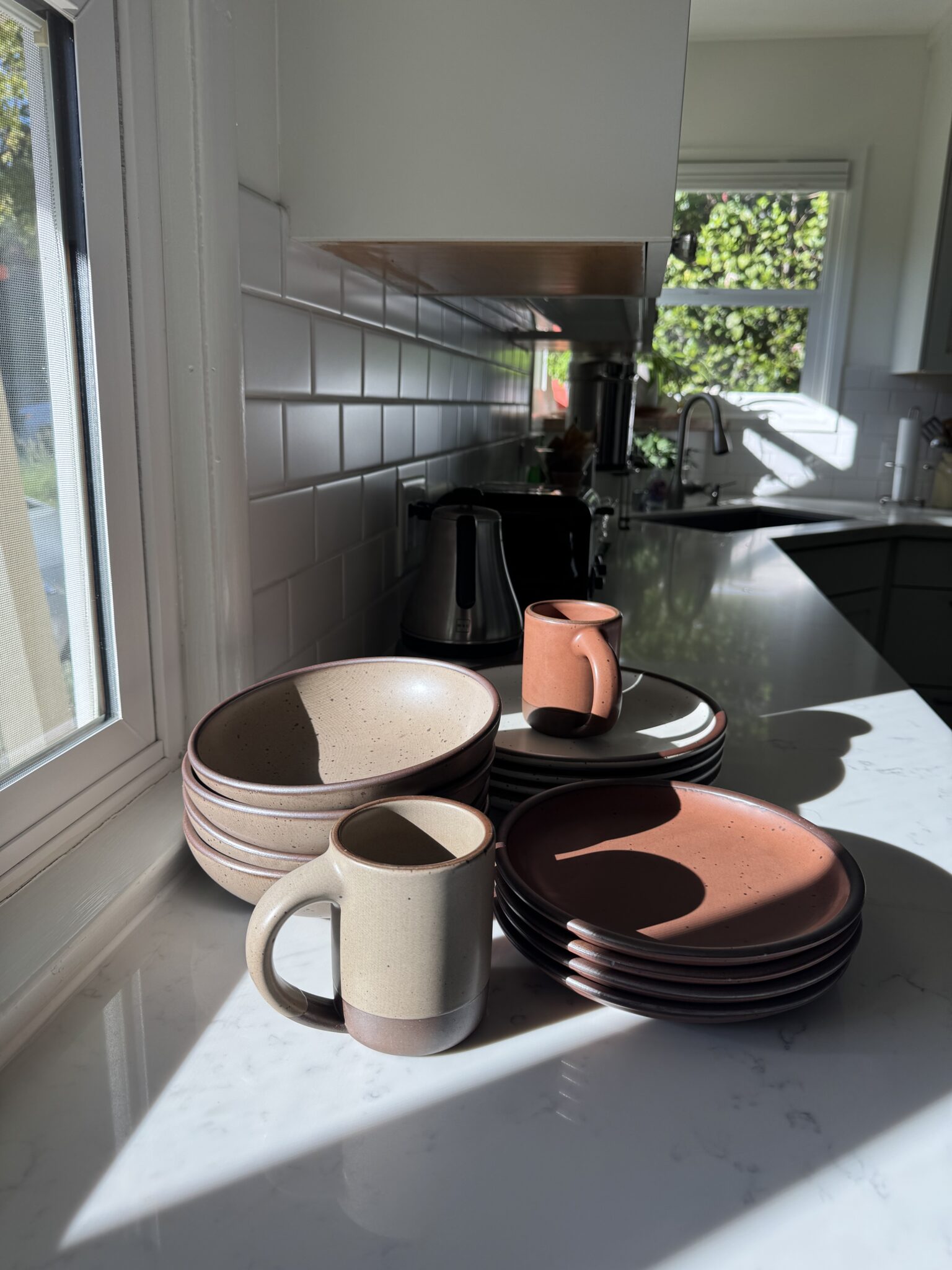 A stack of ceramic dishes and mugs sit on a sunlit kitchen counter near a window, with a sink and appliances visible in the background.