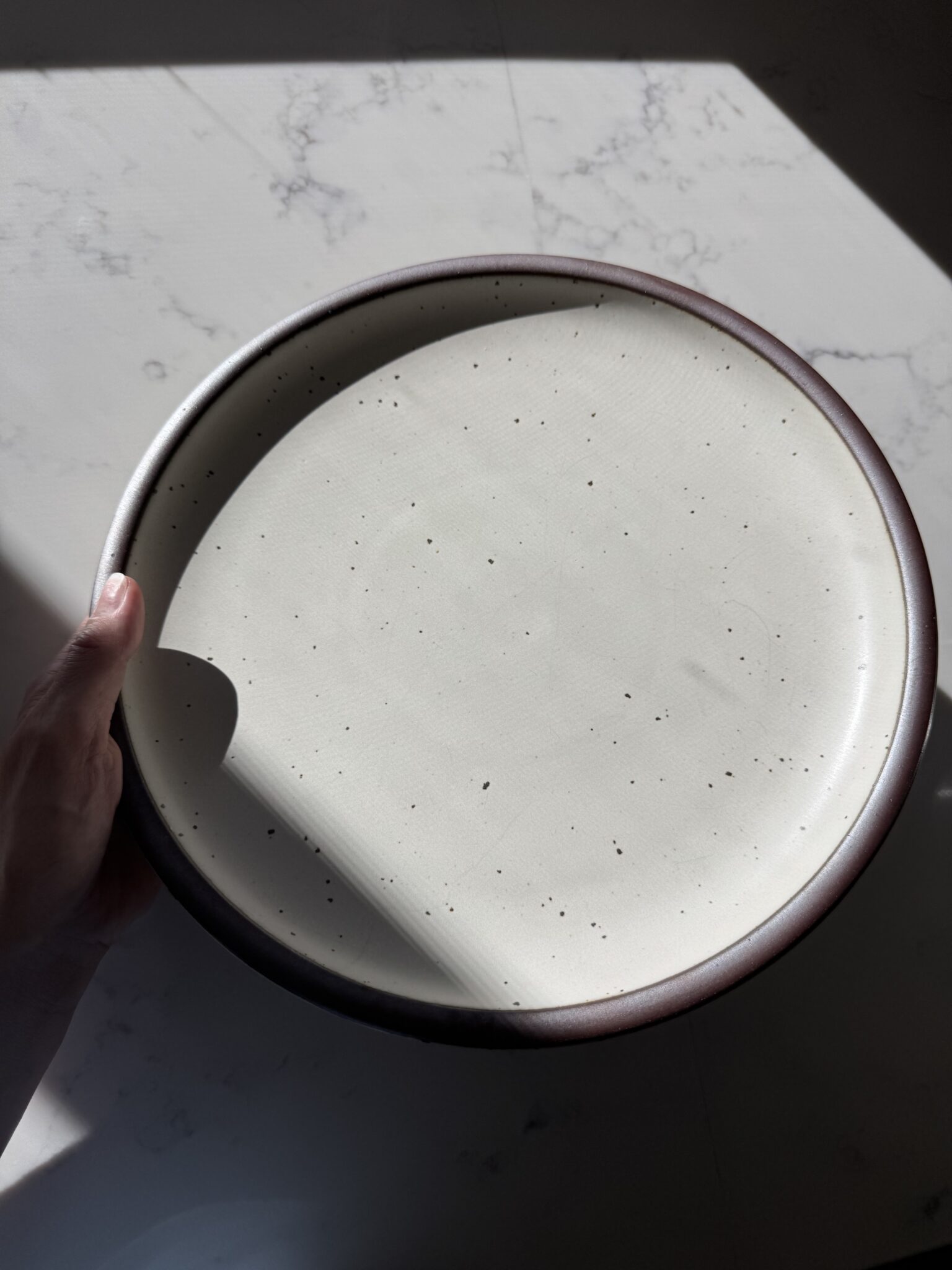 A hand holds a round, cream-colored ceramic plate with a brown rim and dark speckles, in bright sunlight on a light surface.