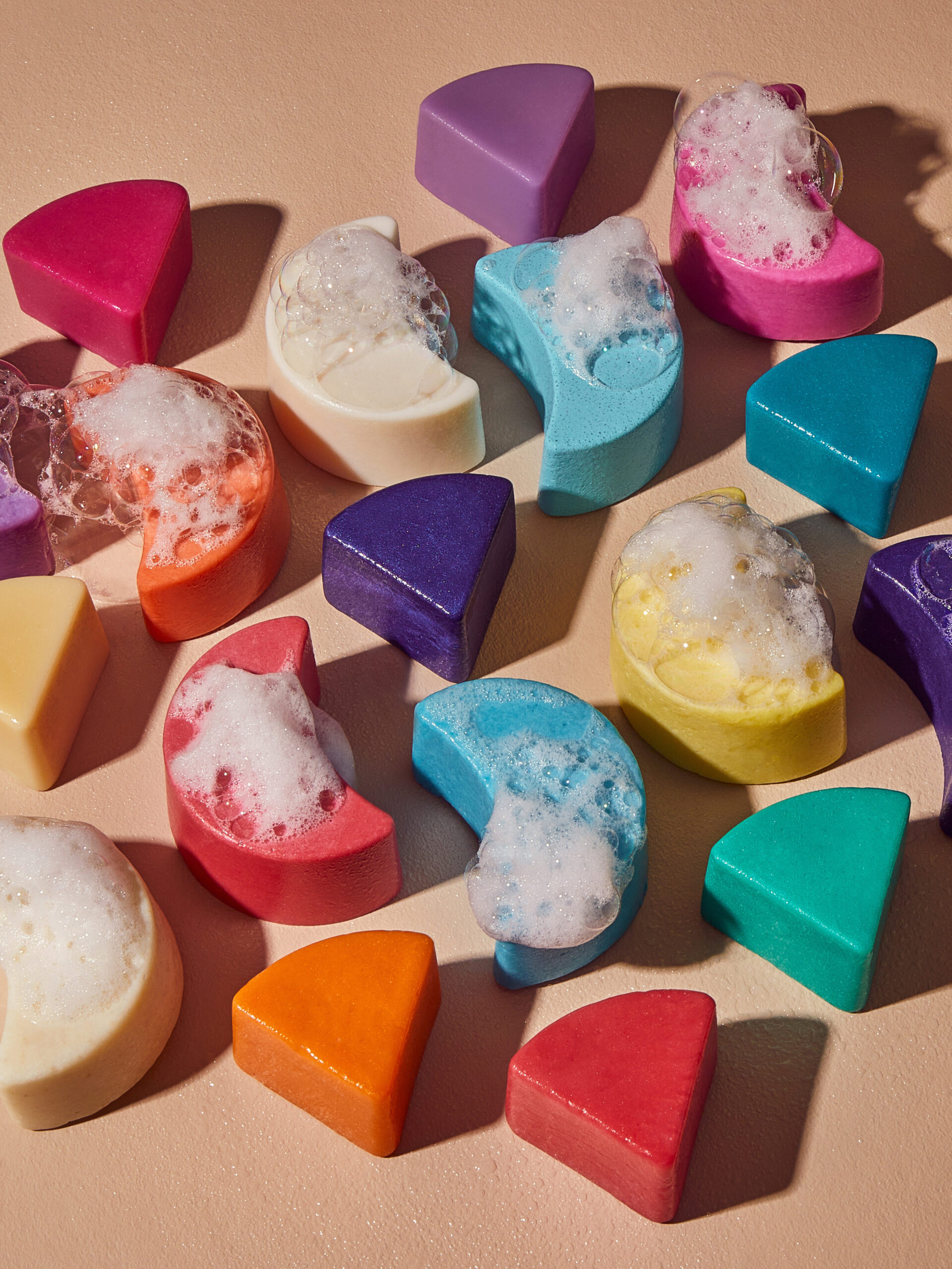 Colorful moon and triangle-shaped soaps, some with bubbles, scattered on a beige surface.