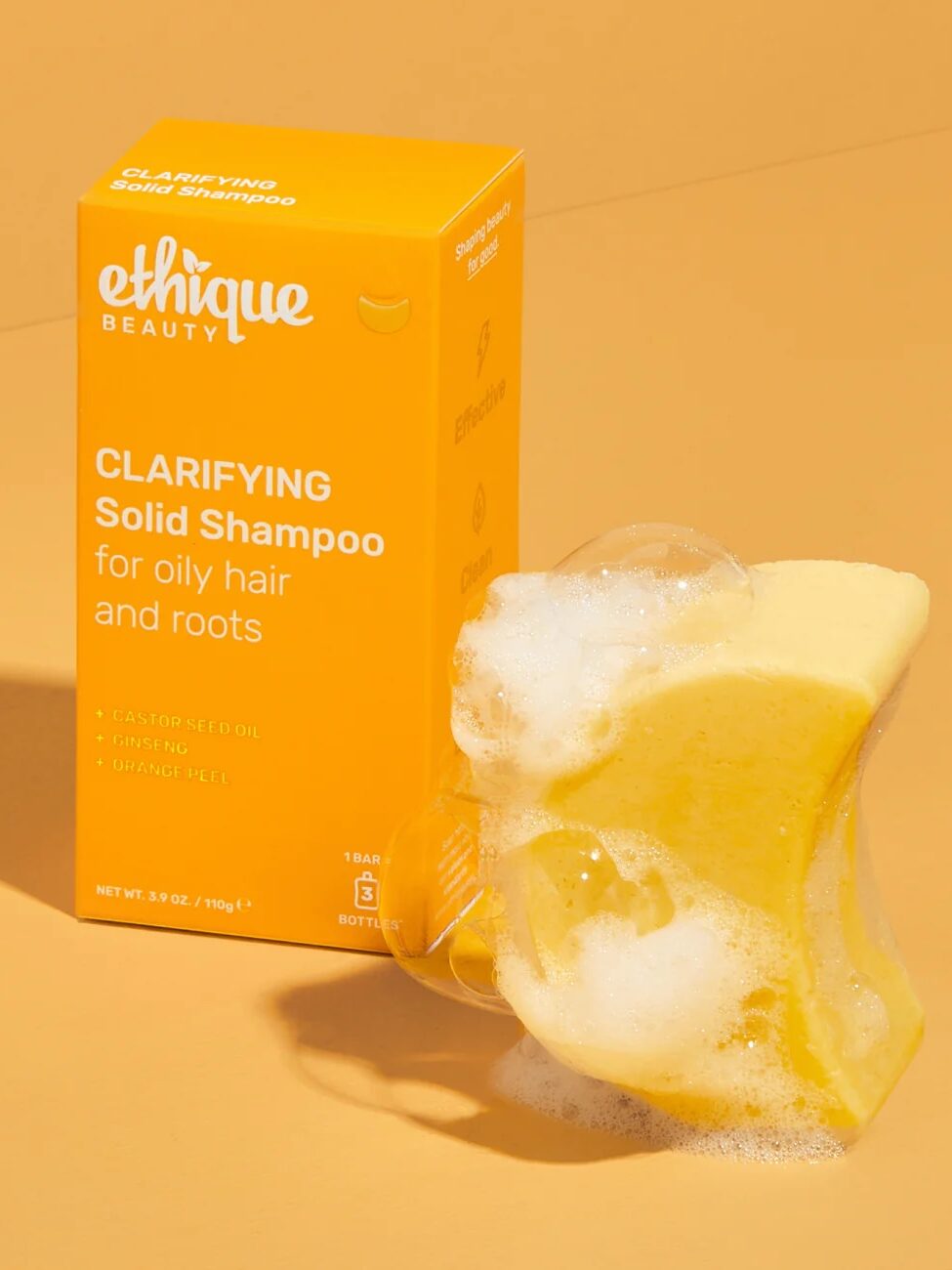 A yellow box labeled "Ethique Clarifying Solid Shampoo" stands next to the shampoo bar, which is foamy and wet, on a solid orange surface.