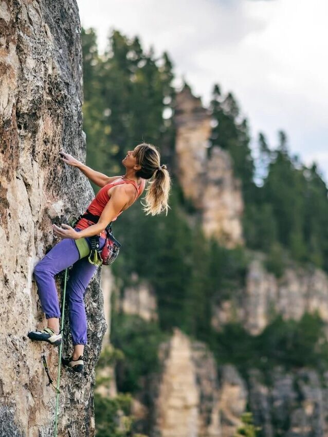 A woman in climbing gear ascends a rocky cliff face outdoors, with trees and cliffs visible in the background.