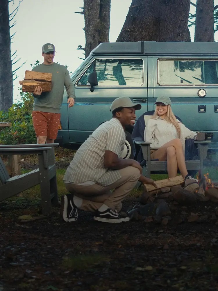 Three people at a campsite; one person carries firewood while two others sit by a campfire near a teal van surrounded by trees.