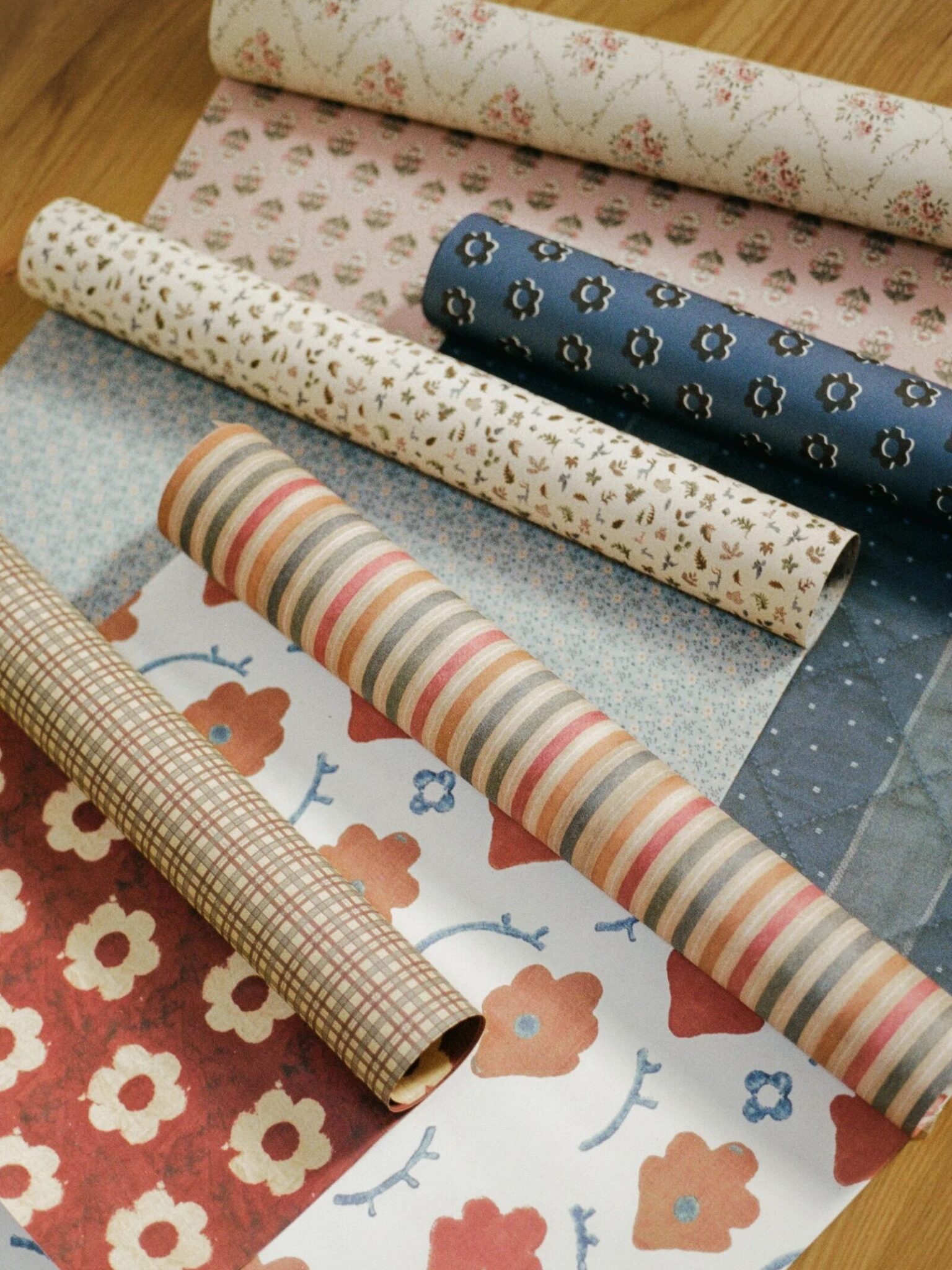 Seven rolls of patterned paper in various colors and floral, plaid, and striped designs are laid out on a wooden surface.