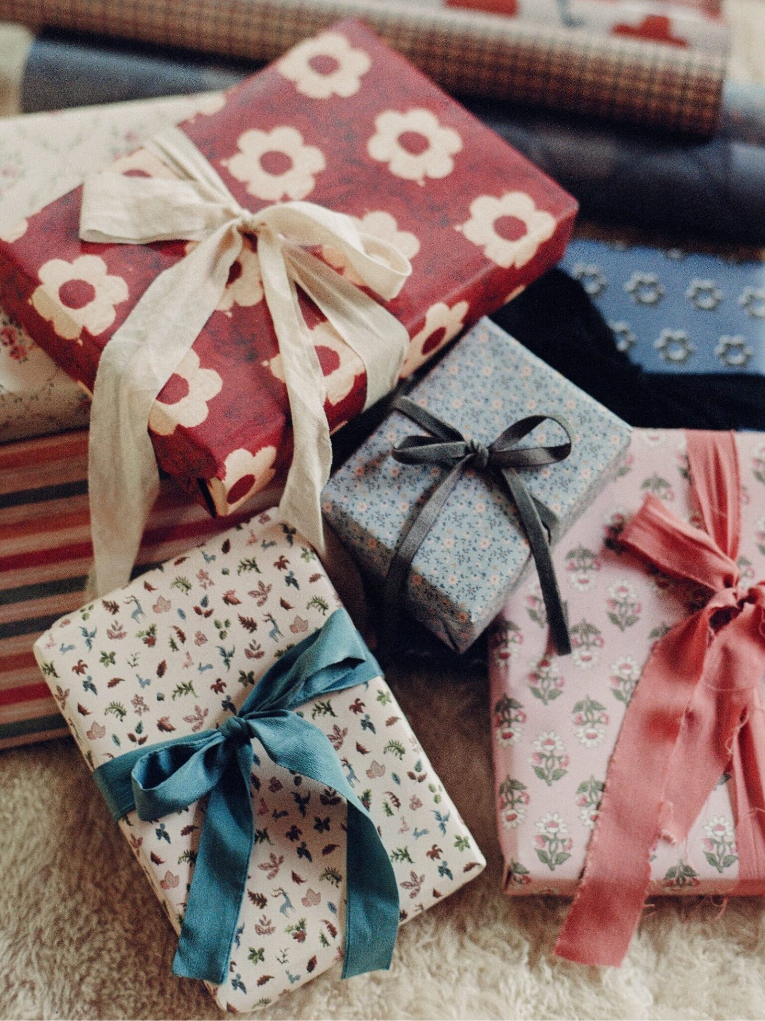 Several wrapped gifts with patterned paper and colorful ribbons are arranged on a soft surface, with extra rolls of wrapping paper in the background.