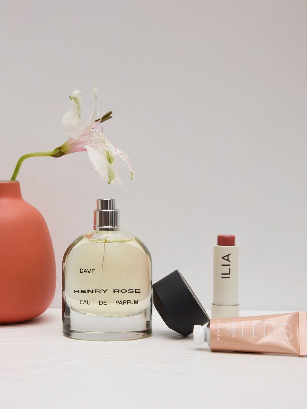 Perfume bottle labeled "Henry Rose," a lipstick, a small tube of makeup, and a pink vase with a white flower on a white surface.