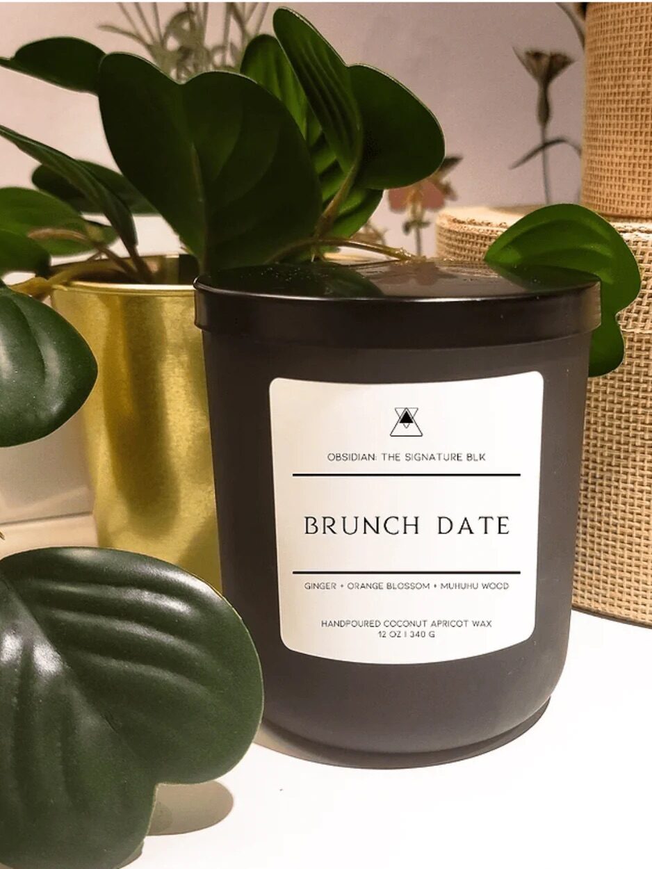 Black candle labeled "Brunch Date" with ginger, orange blossom, and muiruri wood scent, on a table next to green plants.