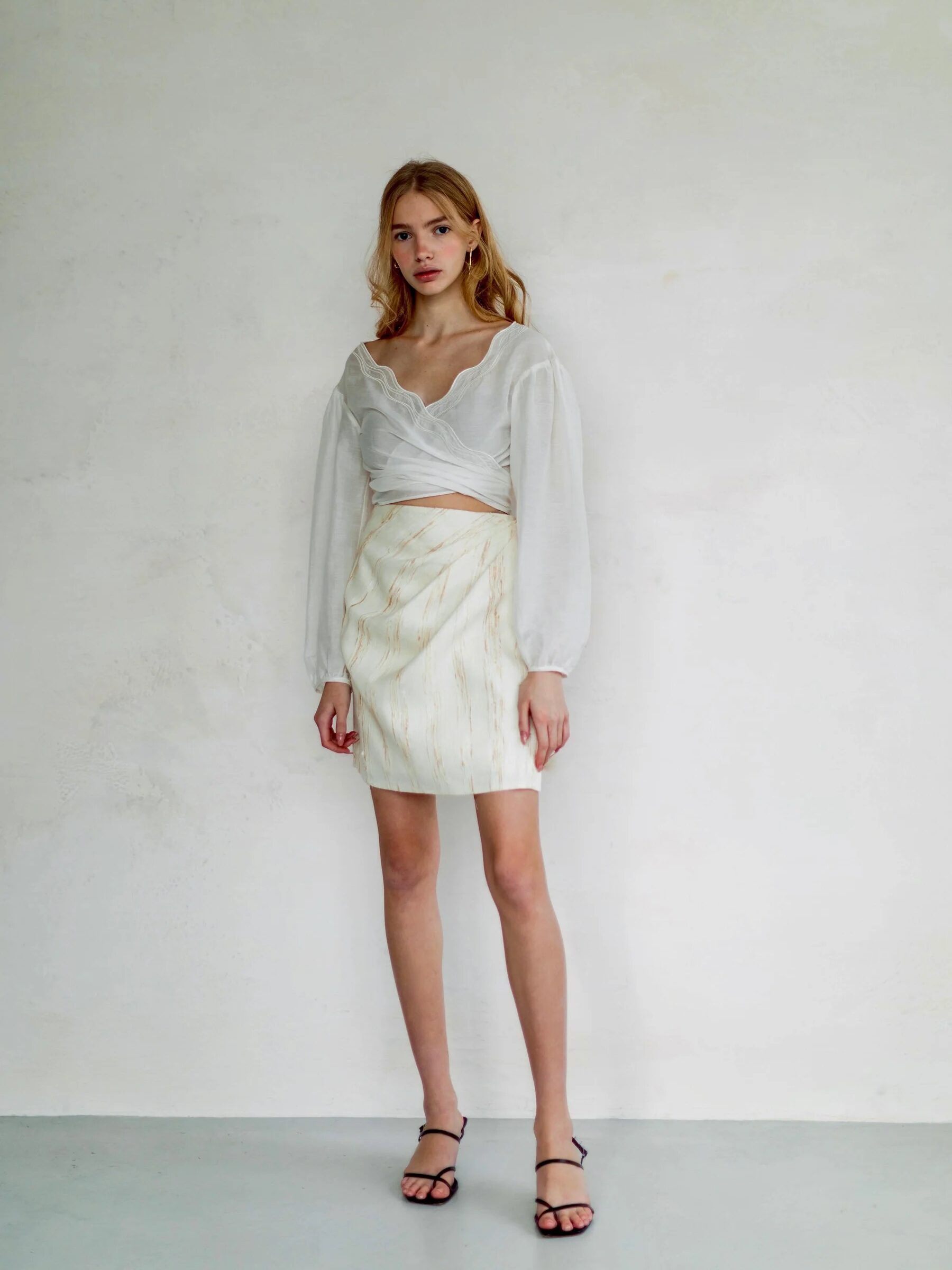 Young woman in a light outfit stands against a plain background, wearing a sheer blouse, white skirt, and black sandals.