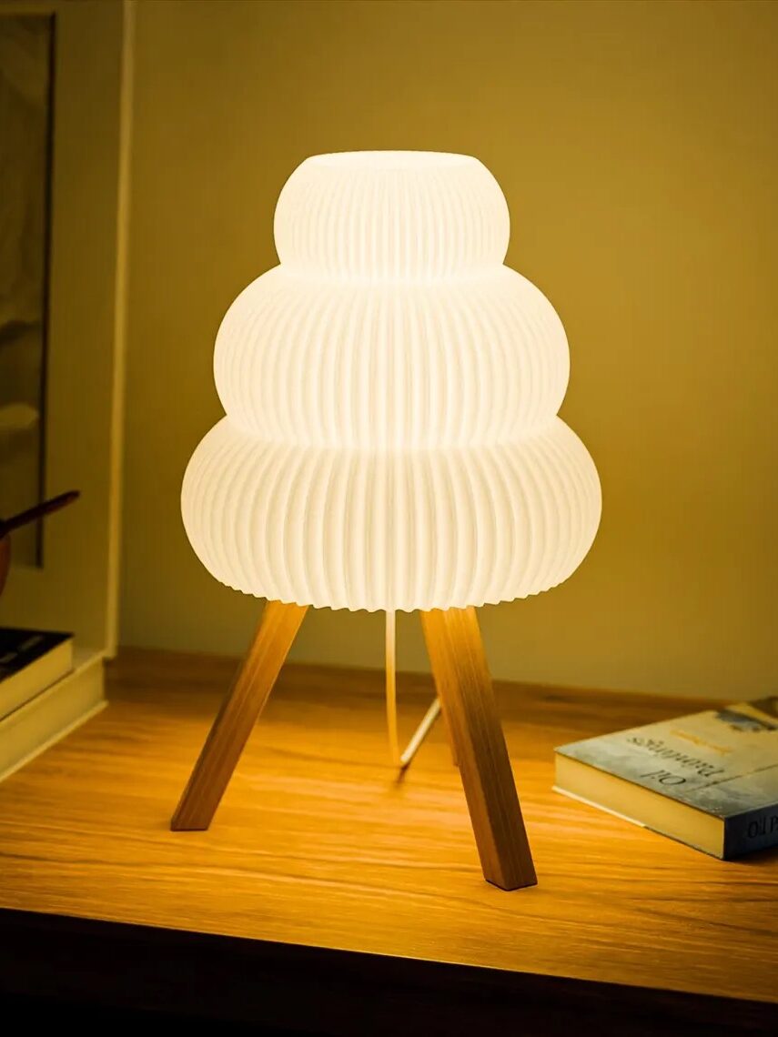 A glowing table lamp with a tiered, pleated white shade on wooden tripod legs is placed on a wooden surface next to a stack of books.