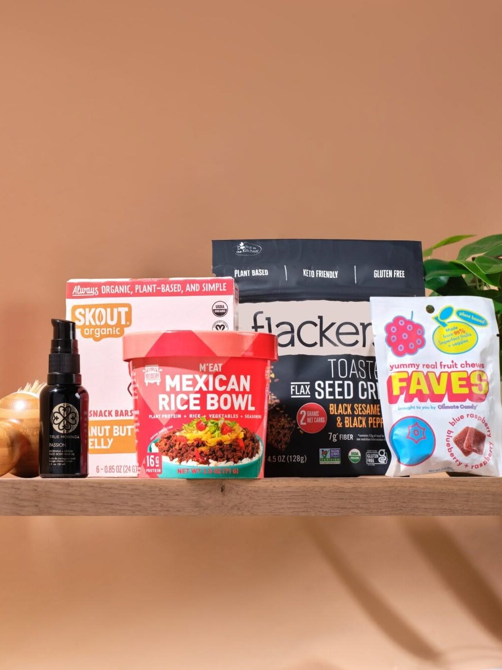 A variety of packaged food items including Mexican rice bowl, seed crackers, fruit chews, organic protein bars, and a bottle of oil displayed on a wooden shelf.