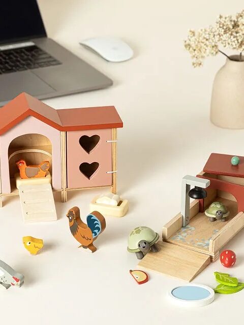 Wooden farm animal toys including a henhouse and feeding area are placed on a desk near a laptop, mouse, and vase with dried flowers.