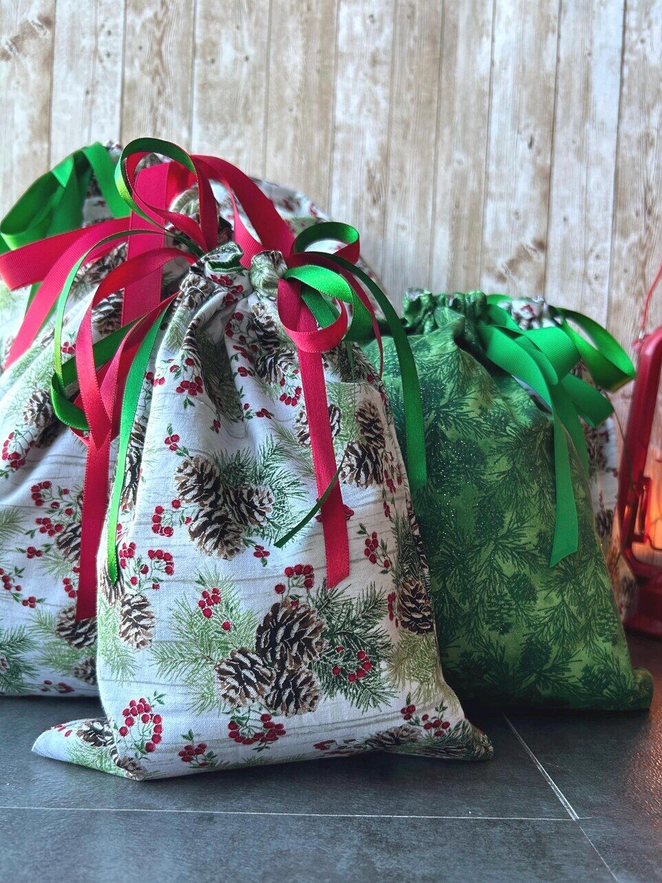 Three fabric gift bags with holiday patterns and ribbons sit on a gray tile floor next to a lit red lantern against a wooden backdrop.