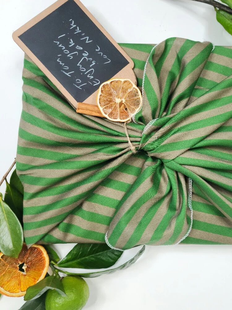 A gift wrapped in green and beige striped fabric, decorated with dried orange slices and green leaves, with a chalkboard gift tag attached.