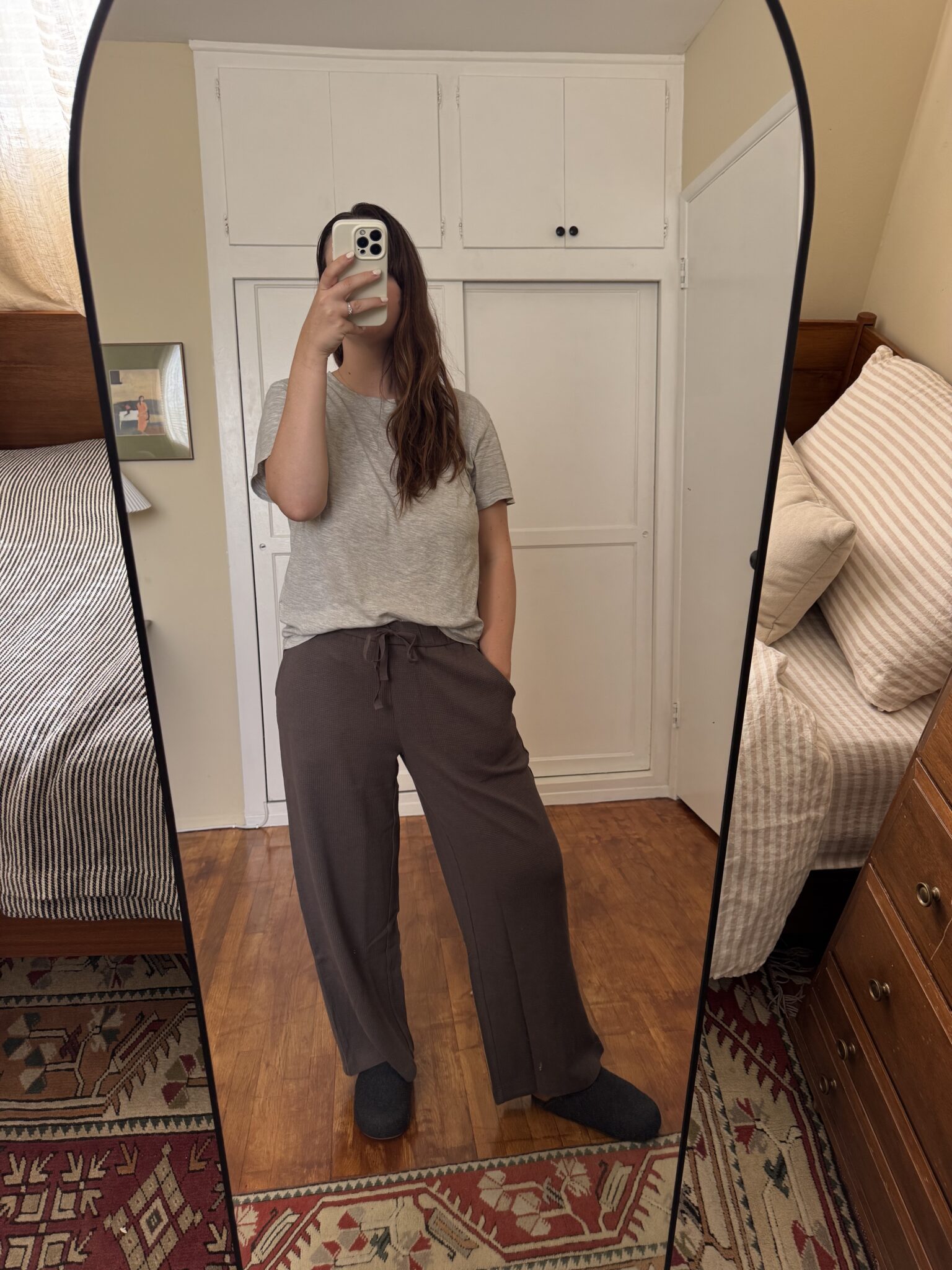 Person taking a mirror selfie in a bedroom, wearing a gray t-shirt, brown drawstring pants, and dark slippers. Bed and dresser are visible in the background.