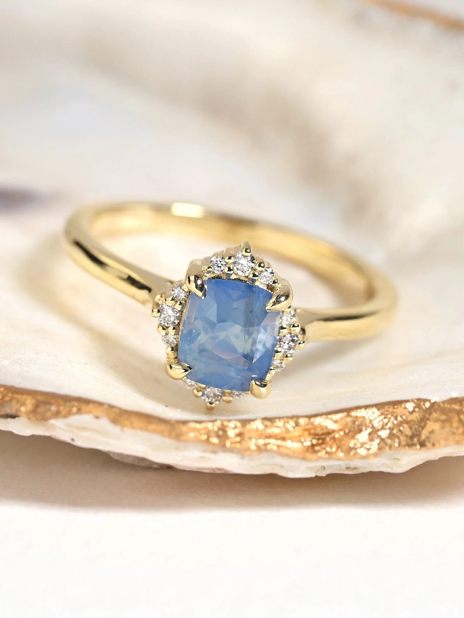 A gold ring with a blue gemstone centered on a textured shell. The gemstone is surrounded by small clear stones.