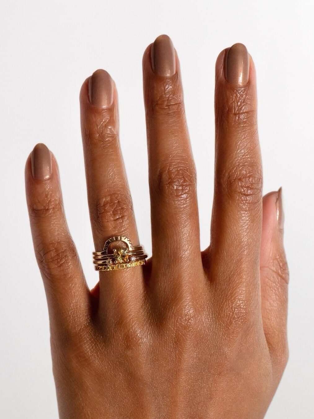 A hand with manicured nails wearing gold rings, including a gemstone ring on the ring finger, is shown against a plain background.